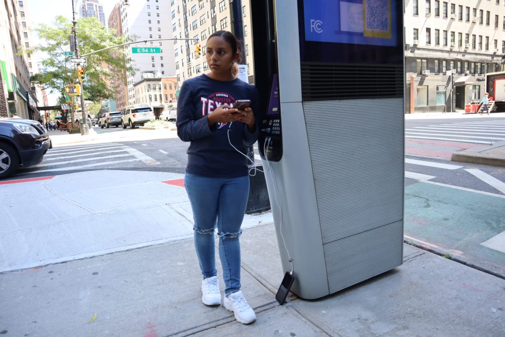 A young woman stands on a New York City street charging her phone on a kiosk
