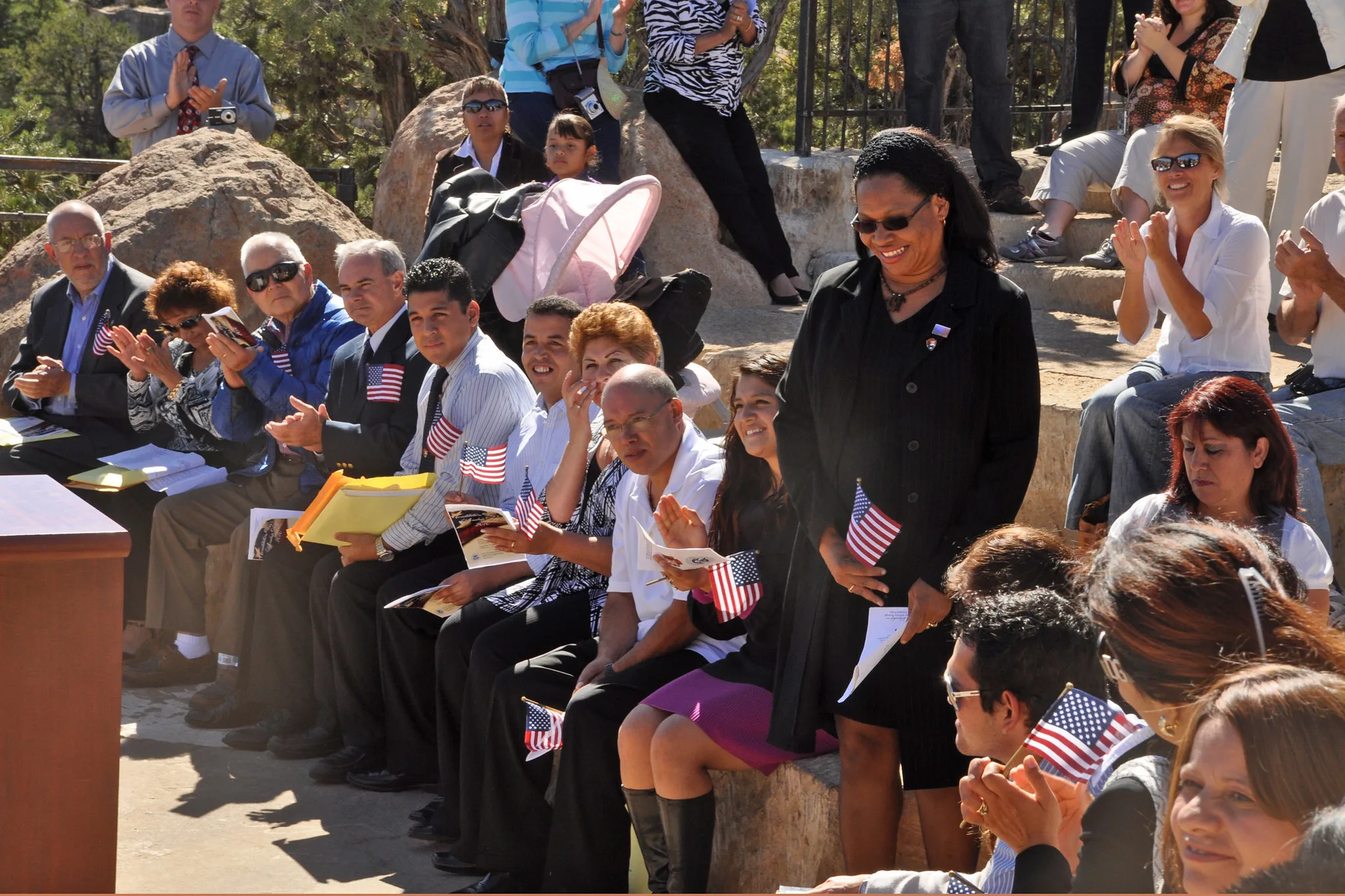 A row of people sit on a stone bench holding small American flags.